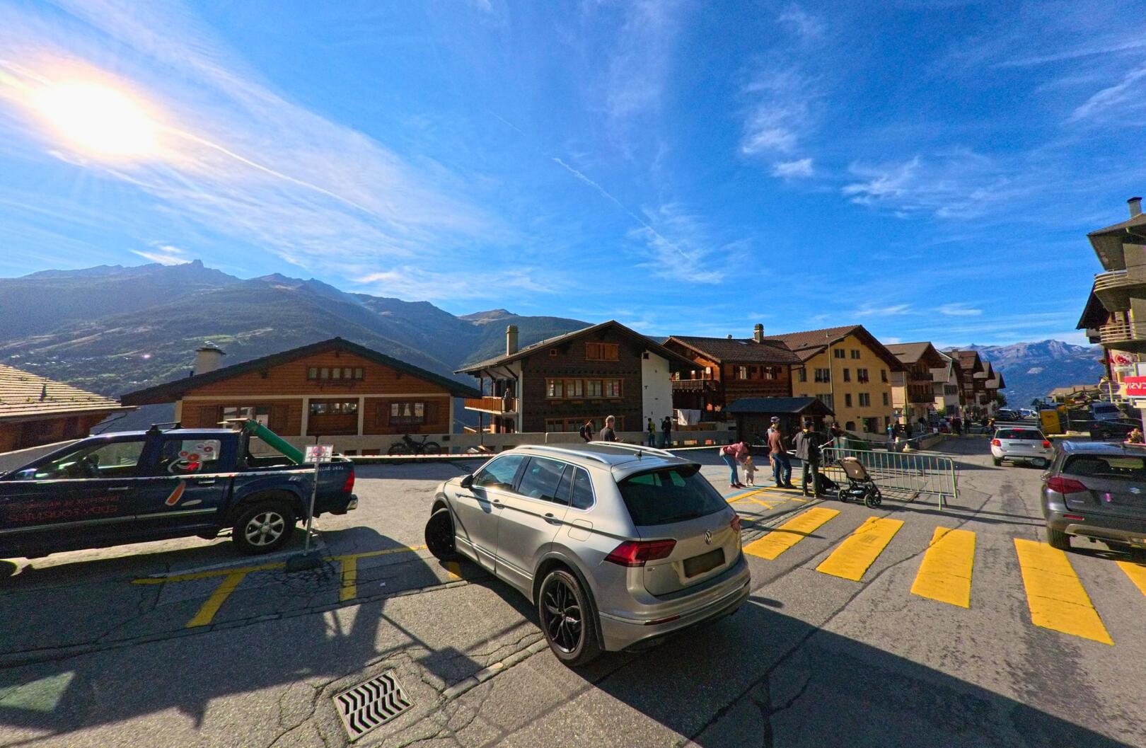 In Hérémence (1987, Kanton Wallis) überqueren Menschen einen Zebrastreifen im Dorfzentrum, während im Hintergrund traditionelle Holzhäuser und Berge sichtbar sind.