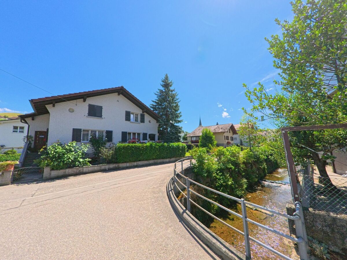  In Hergiswil bei Willisau (6133, Kanton Luzern) verläuft eine schmale Strasse entlang eines Bachs, gesäumt von Wohnhäusern und grüner Bepflanzung mit Blick auf den Kirchturm im Hintergrund.