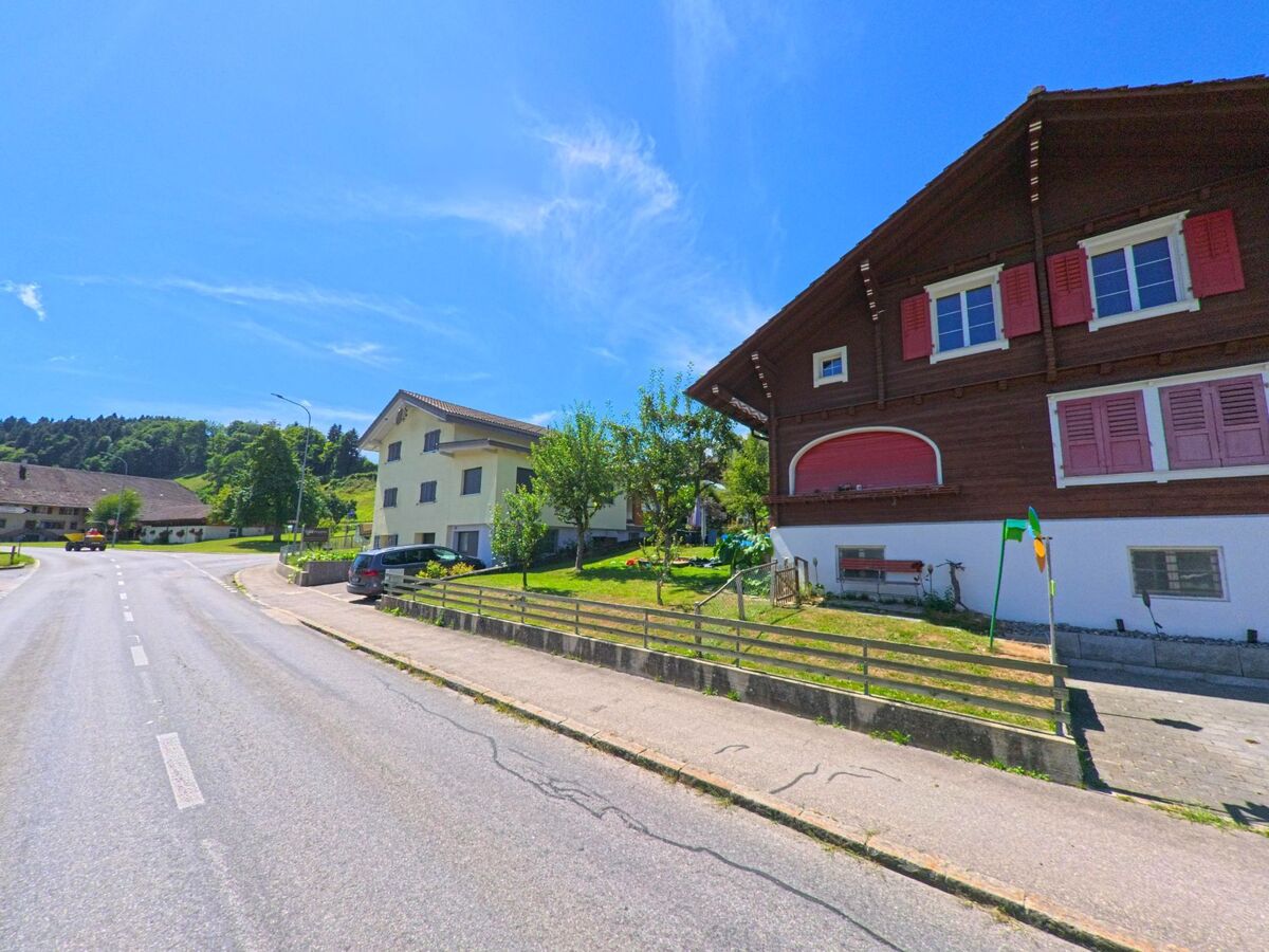 In Hergiswil bei Willisau (6133, Kanton Luzern) stehen ein braunes Chalet mit roten Fensterläden und weitere Wohnhäuser entlang einer ruhig verlaufenden Dorfstrasse.