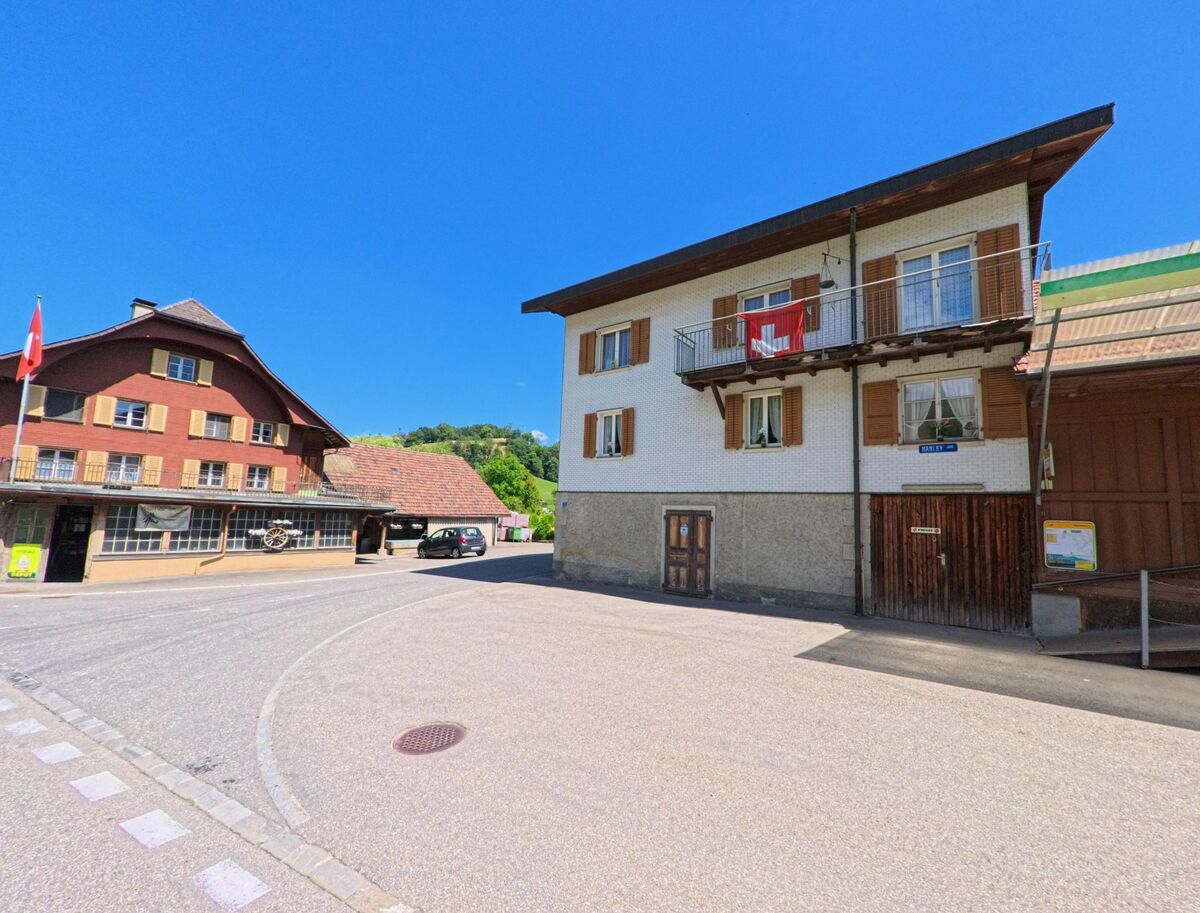  Im Dorfzentrum von Hergiswil bei Willisau (6133, Kanton Luzern) stehen traditionelle Gebäude mit Balkon und Schweizer Flagge neben einem Landi-Geschäft und einem historischen Gasthaus.