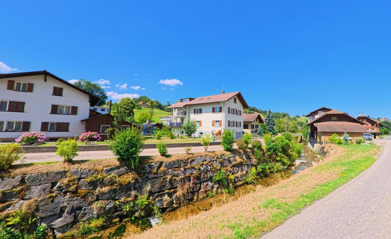 In Hergiswil bei Willisau (6133, Kanton Luzern) verlaufen Wohnhäuser entlang eines kleinen Bachlaufs mit gemauerter Uferbefestigung inmitten grüner Hügellandschaft.