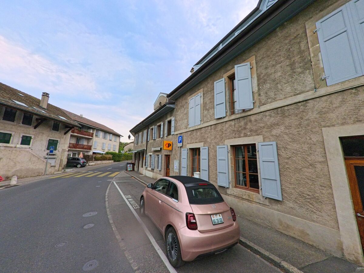 Ein rosafarbenes Auto parkt in Hermance (1248, Kanton Genf) vor einem Haus mit hellblauen Fensterläden entlang einer engen Strasse.
