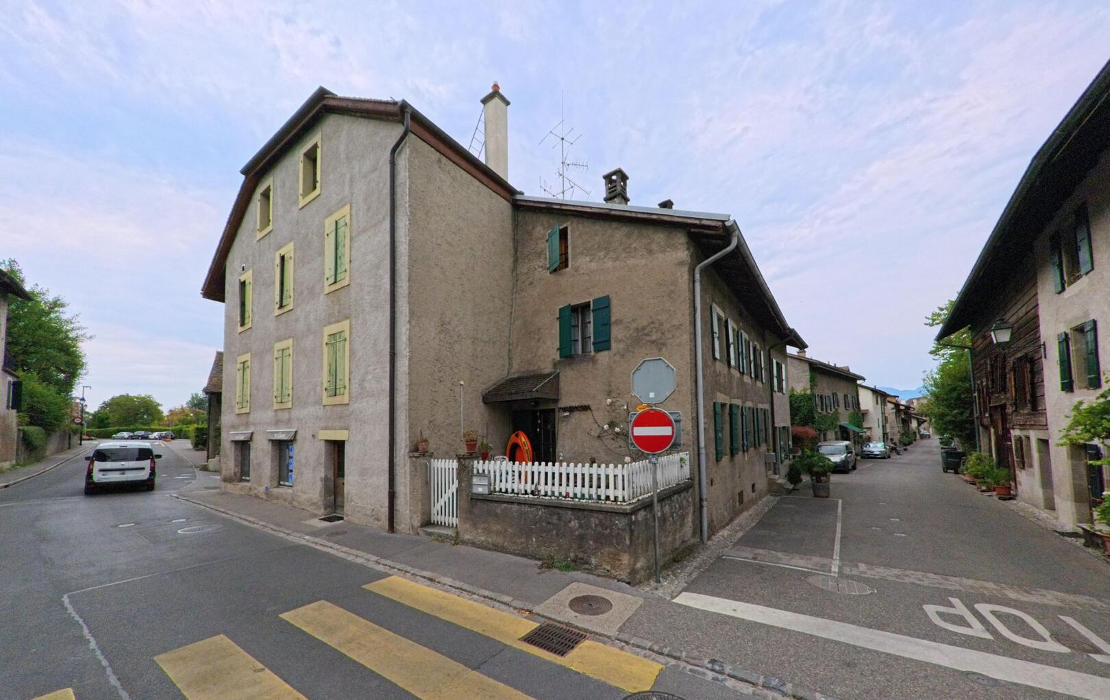 In Hermance (1248, Kanton Genf) steht ein hohes Haus mit hellgrünen Fensterläden an einer Kreuzung mit Fussgängerstreifen.