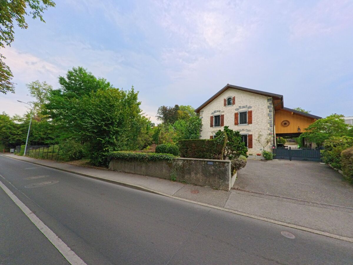Ein renoviertes Wohnhaus mit Holzfassade und Fensterläden steht in Hermance (1248, Kanton Genf) an einer Strasse neben Bäumen.