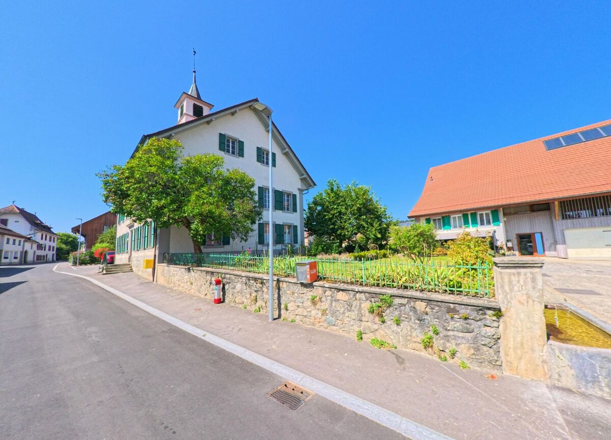 Das Gemeindehaus mit kleinem Turm und gruenen Fensterlaeden steht in Hermenches (1513, Kanton Waadt) an einer Strasse, davor befindet sich ein Garten mit Steinmauer.