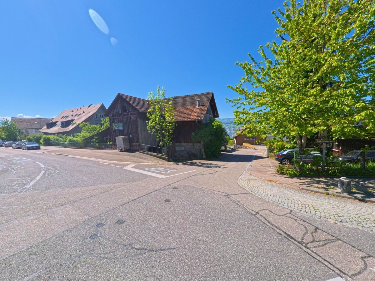 Im Ortskern von Herrliberg, PLZ 8704 im Kanton Zürich, treffen sich mehrere Strassen an einem historischen Holzgebäude, umgeben von Bäumen und geparkten Autos.