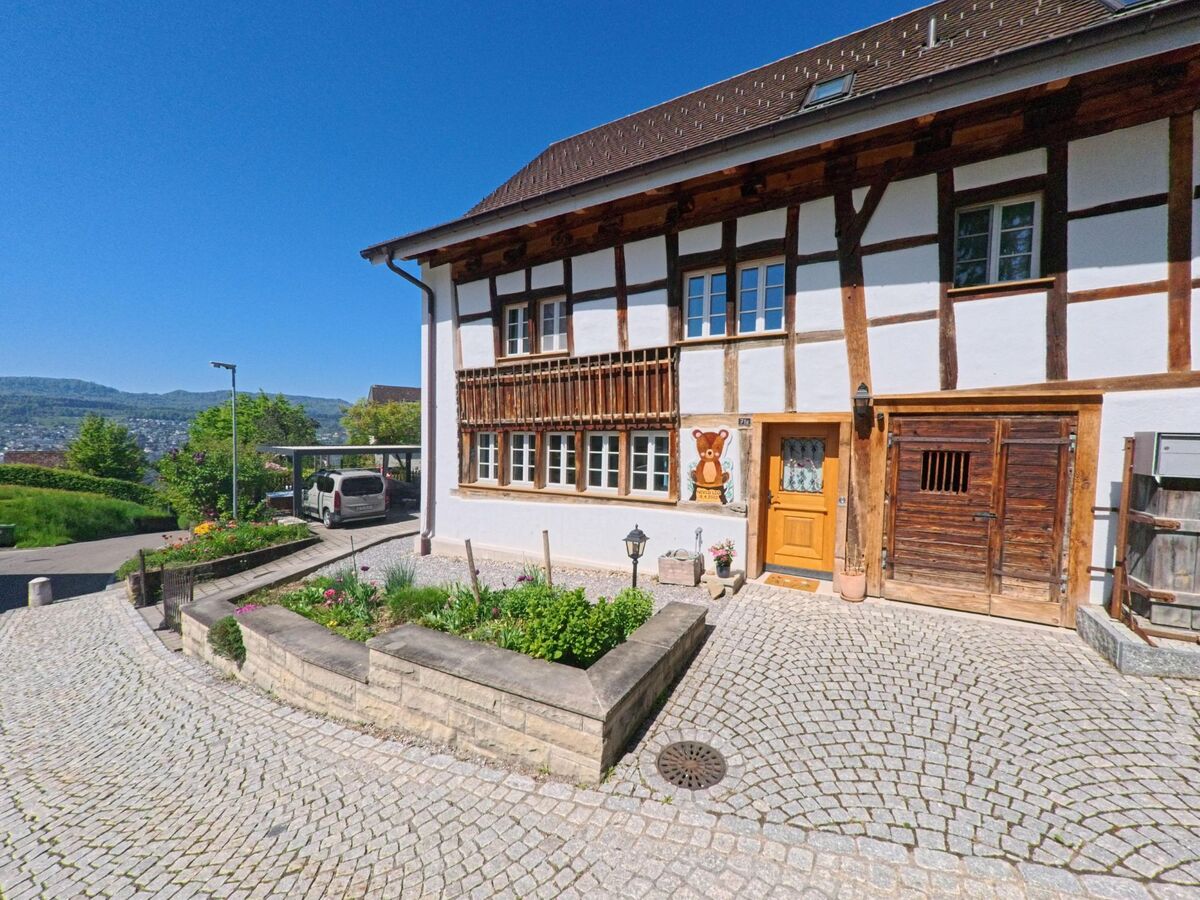 Ein gepflegtes Fachwerkhaus mit Blumenvorgarten steht in Herrliberg im Kanton Zürich, PLZ 8704, oberhalb einer kurvigen Pflasterstrasse mit Ausblick auf das gegenüberliegende Seeufer.