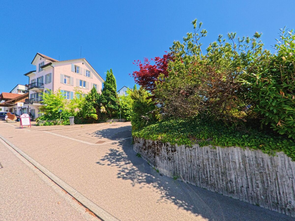 In Herrliberg im Kanton Zürich, PLZ 8704, steht ein helles Mehrfamilienhaus mit Fensterläden an einer sanft ansteigenden Strasse, umgeben von dichter Bepflanzung.