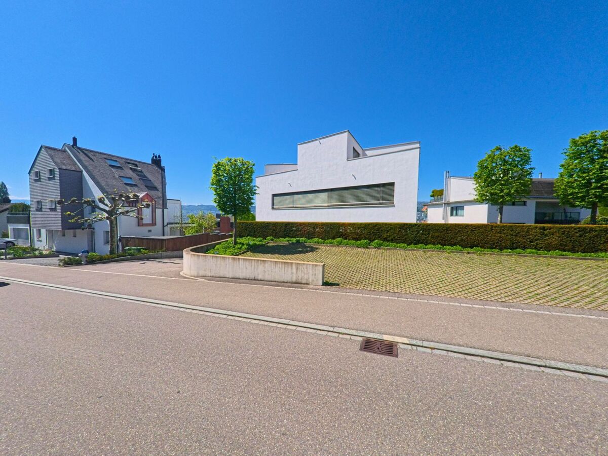 In Herrliberg, PLZ 8704 im Kanton Zürich, stehen moderne Einfamilienhäuser mit klaren geometrischen Formen entlang einer ruhigen Strasse mit begrünter Böschung.