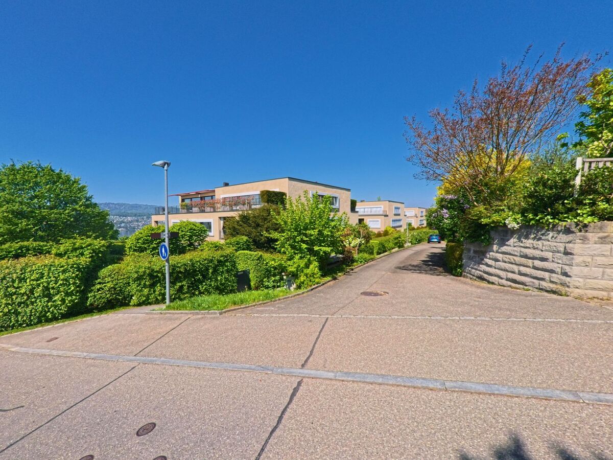 In Herrliberg, PLZ 8704 im Kanton Zürich, verläuft ein schmaler Weg zwischen modernen Wohnhäusern mit begrünten Vorgärten und Blick auf den Hang gegenüber.