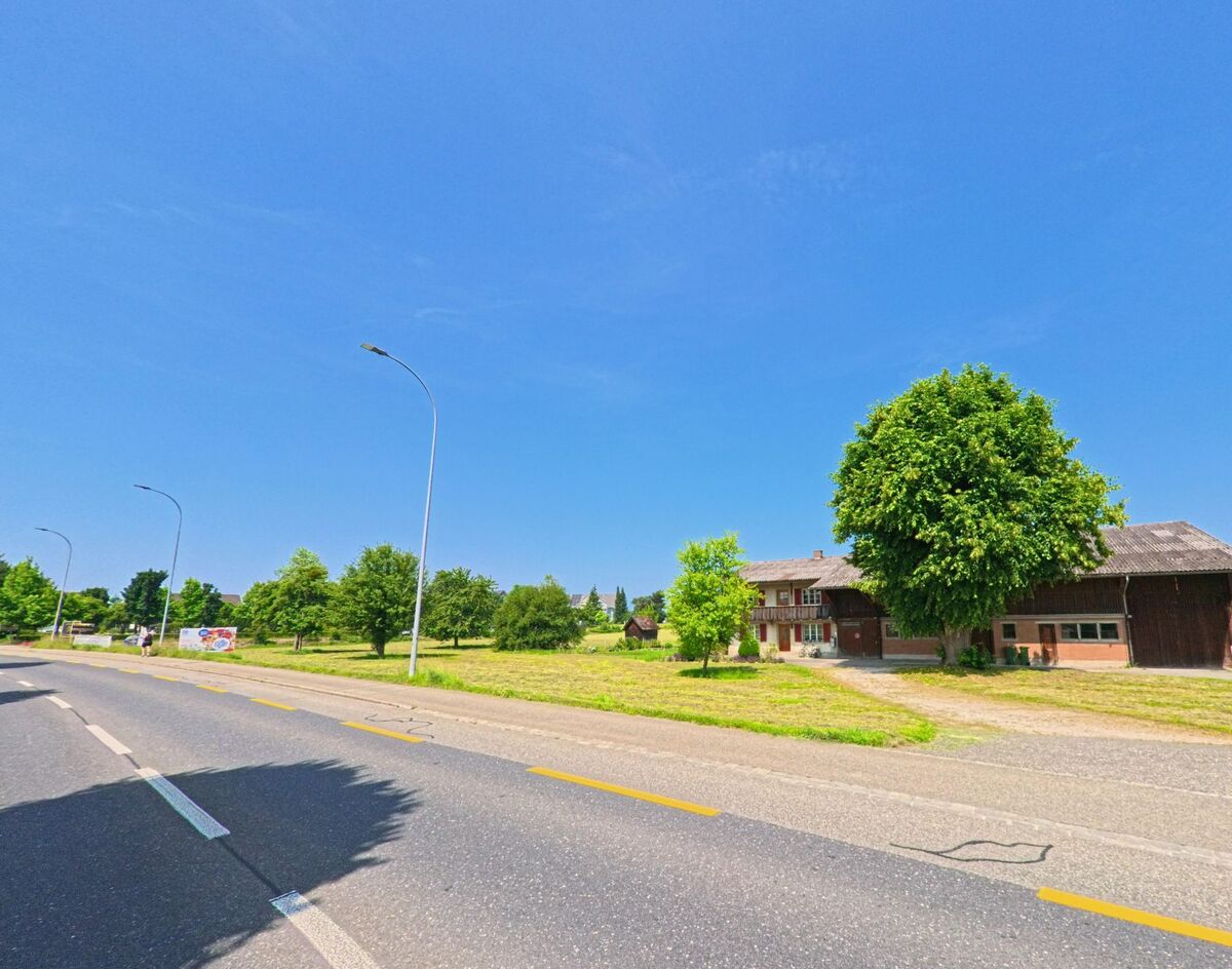 Ein Bauernhof in Herzogenbuchsee (3360, Kanton Bern) steht am Rand einer Straße, umgeben von Bäumen und Feldern, mit einem Lkw, der auf einem nahegelegenen Weg geparkt ist.