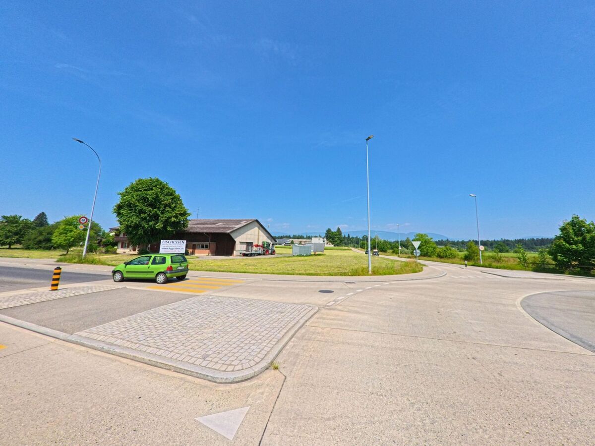 Ein Kreisverkehr in Herzogenbuchsee (3360, Kanton Bern) mit einem grünen Auto, das den Kreisverkehr verlässt, und einem modernen Gebäude im Hintergrund.