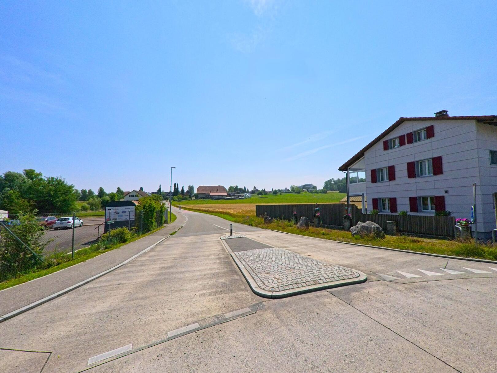 In Herzogenbuchsee (3360, Kanton Bern) führt eine ruhige Strasse an einem modernen Gebäude entlang, mit einem weiten Blick auf Felder und eine Parkfläche im Hintergrund.