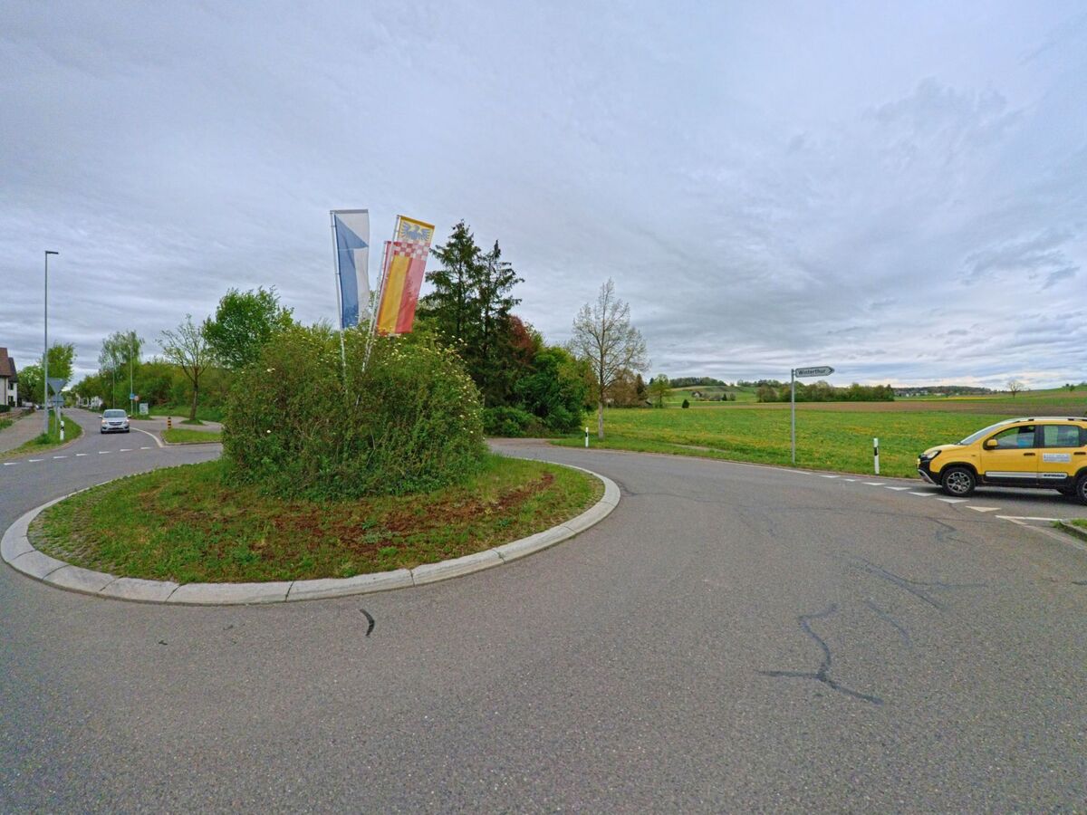 Ein Kreisverkehr mit beflaggtem Mittelbeet markiert in Hettlingen im Kanton Zürich (PLZ 8442) den Übergang vom Siedlungsgebiet zu einer offenen Feldlandschaft.