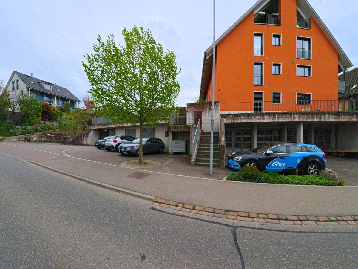 In Hettlingen im PLZ-Gebiet 8442 im Kanton Zürich stehen ein modernes orangefarbenes Mehrfamilienhaus und mehrere abgestellte Fahrzeuge entlang einer Quartierstrasse.