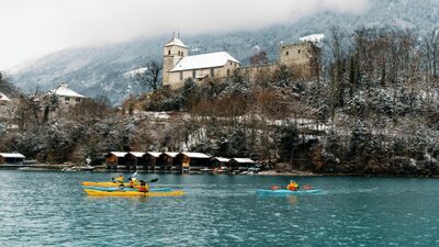 Auf dem türkisblauen Brienzersee bei Bönigen (3806, Kanton Bern) paddeln Kajakfahrer der Hightide Kayak School vor verschneiten Uferhängen und einer historischen Kirche.