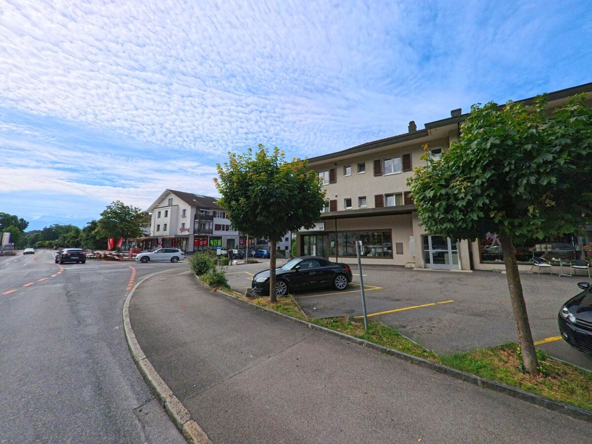 In Hilterfingen (3626, Kanton Bern) säumen moderne Wohn- und Geschäftshäuser mit Parkplätzen eine breite Hauptstrasse mit Blick auf die Berge.