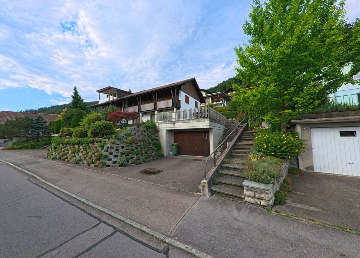 In Hilterfingen (3626, Kanton Bern) steht ein helles Einfamilienhaus mit grünen Fensterläden und gepflegtem Garten am Fusse eines bewaldeten Hangs.