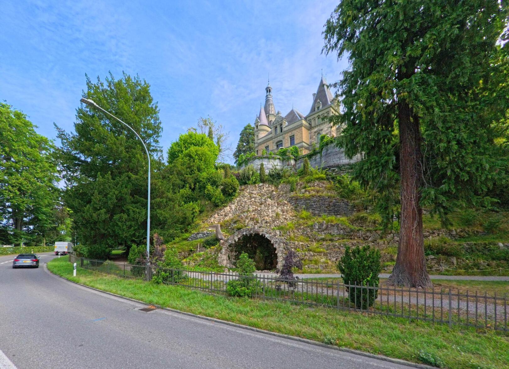 In Hilterfingen (3652, Kanton Bern) führt eine kurvige Strasse an einem bewaldeten Hang vorbei, auf dem ein historisches Schloss mit Türmen thront.