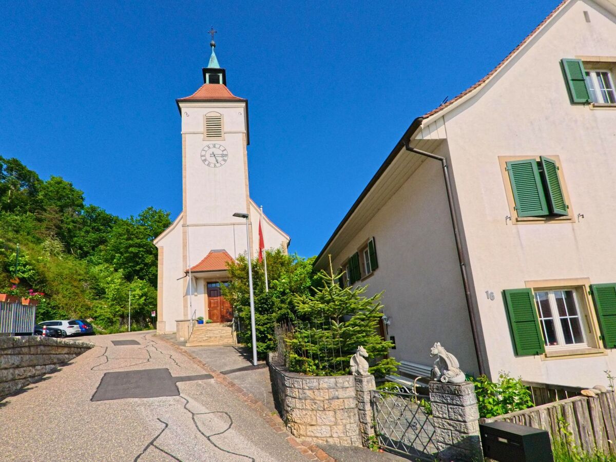 In Himmelried (4204, Kanton Solothurn) erhebt sich der helle Kirchturm mit Uhr über der ansteigenden Strasse, flankiert von Wohnhäusern und Bäumen.