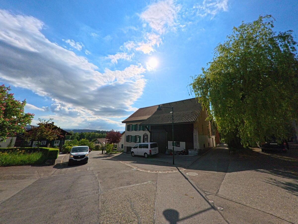 In Himmelried (4204, Kanton Solothurn) fällt die Nachmittagssonne auf eine ruhige Kreuzung mit Wohnhäusern, Bäumen und geparkten Fahrzeugen.