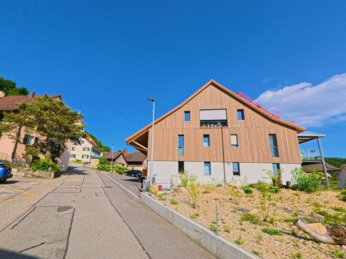 In Himmelried (PLZ 4204, Kanton Solothurn) verläuft eine leicht ansteigende Strasse zwischen traditionellen und modernen Wohnhäusern mit Sicht auf den blauen Himmel.