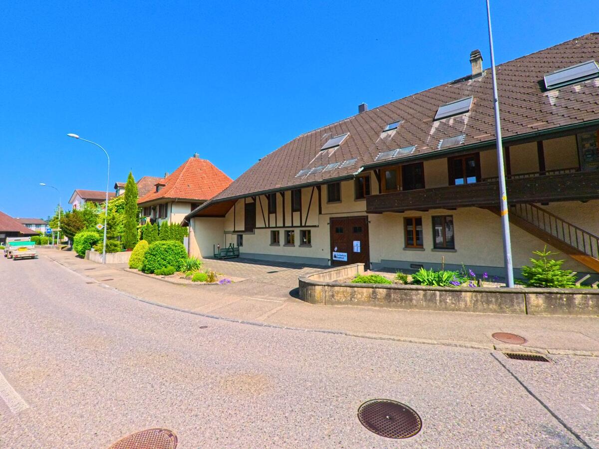 In Hindelbank (3324, Kanton Bern) säumen traditionelle Wohnhäuser mit Satteldächern und gepflegten Vorgärten eine leicht befahrene Strasse bei strahlendem Wetter.