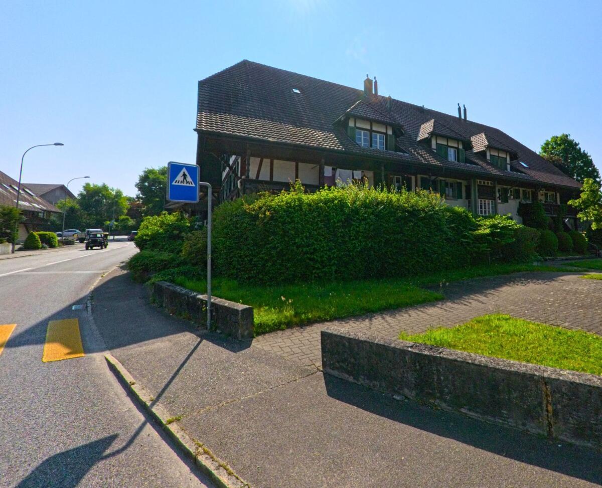 In Hindelbank (3324, Kanton Bern) steht ein traditionelles Gebäude mit dunklem Schindeldach und Fachwerkfassade gegenüber einem kleinen Brunnen am Strassenrand.