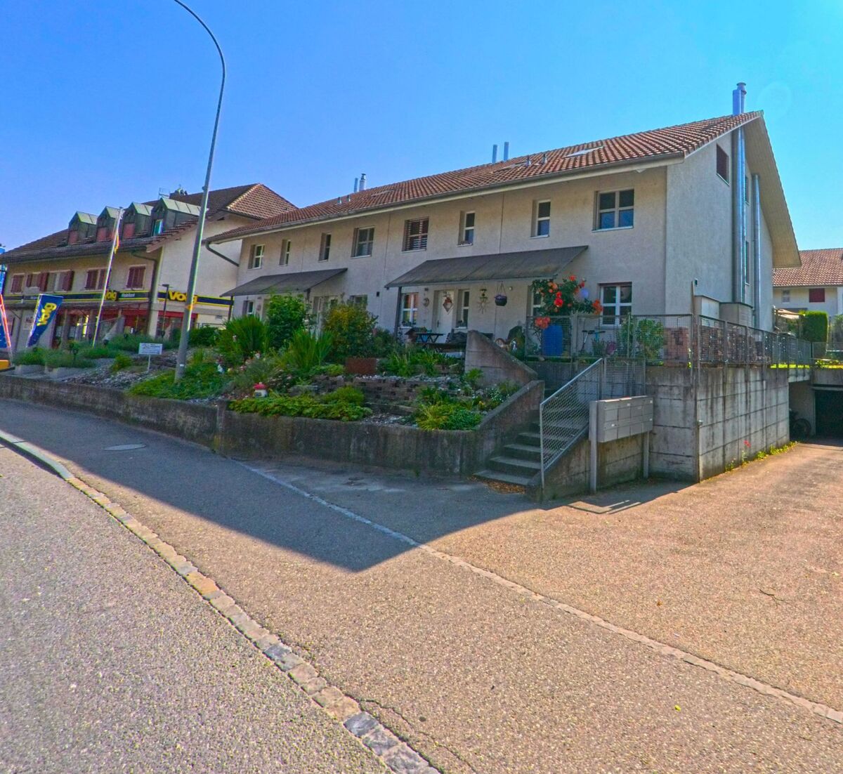 In Hindelbank (3324, Kanton Bern) stehen Reihenhäuser mit kleinen Vorgärten und separatem Garagenzugang an einer leicht gebogenen Strasse bei hellem Sonnenschein.