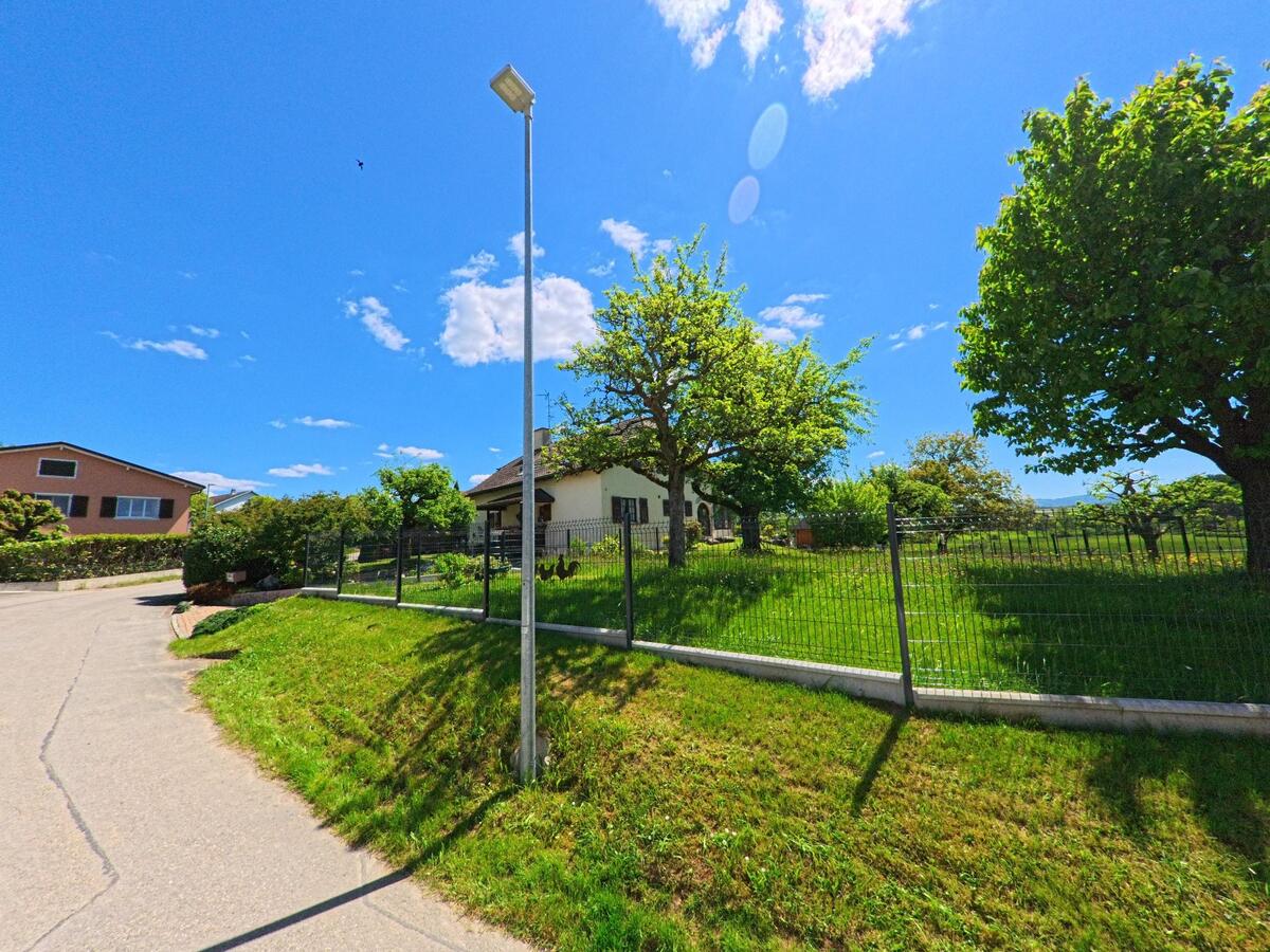 Hinter einem Metallzaun liegt in Damphreux (2933, Kanton Jura) ein gepflegter Garten mit mehreren Bäumen und einem Wohnhaus unter blauem Himmel..jpg