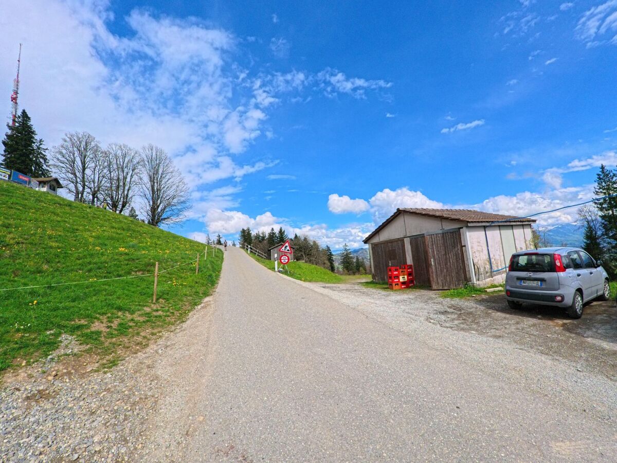 In Hinwil im Kanton Zürich (PLZ 8342) steigt ein schmaler Weg auf eine Anhöhe mit Sicht auf einen Sendeturm an, vorbei an einer Hütte und parkenden Fahrzeugen.