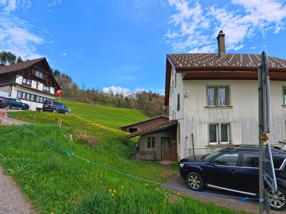 In Hinwil im Kanton Zürich (PLZ 8342) stehen zwei Wohnhäuser am Hang, umgeben von Wiesen und Fahrzeugen, mit einer kleinen Zufahrt und Blick in bewaldetes Gelände.