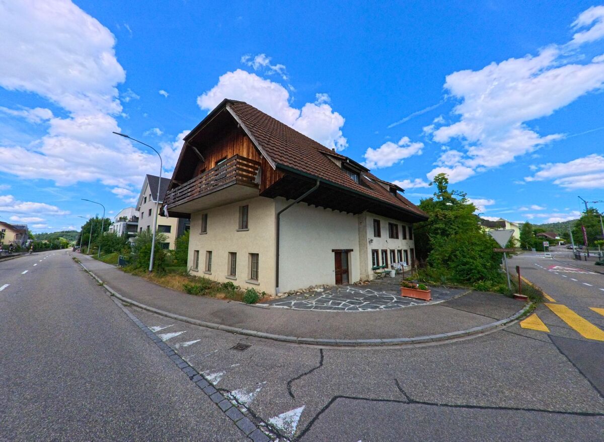 An einer Strassenkreuzung in Hirschthal (5042, Kanton Aargau) steht ein traditionelles Wohnhaus mit Holzbalkon und Krueppelwalmdach neben modernen Nachbargebaeuden.