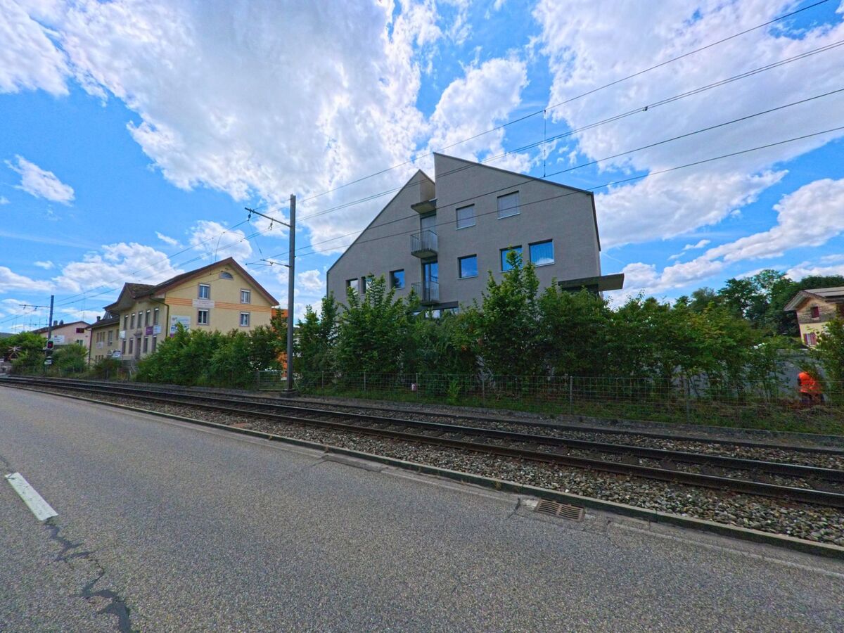 In Hirschthal (5042, Kanton Aargau) verlaufen Bahngeleise parallel zu einer Strasse mit Wohnhaeusern, darunter ein modernes Mehrfamilienhaus mit markanter Dachform.