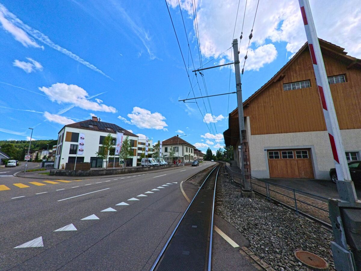 In Hirschthal (5042, Kanton Aargau) verlaeuft eine Bahnlinie unmittelbar neben einer Hauptstrasse, flankiert von einem modernen Wohnhaus und einer grossen Scheune mit Holzelementen.
