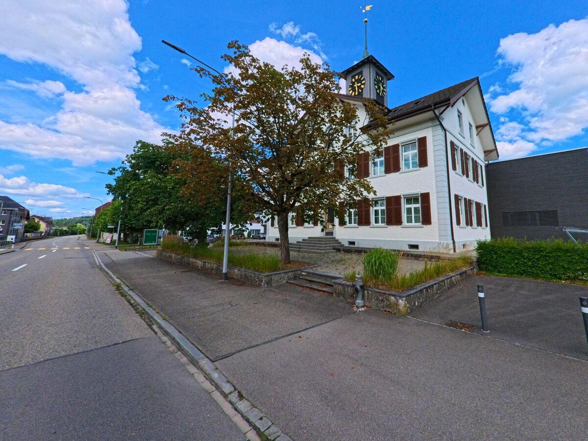 Das Gemeindehaus von Hirschthal (5042, Kanton Aargau) mit Turmuhr und Fensterlaeden steht an einer Hauptstrasse, flankiert von einem Baum und einem kleinen Vorplatz.