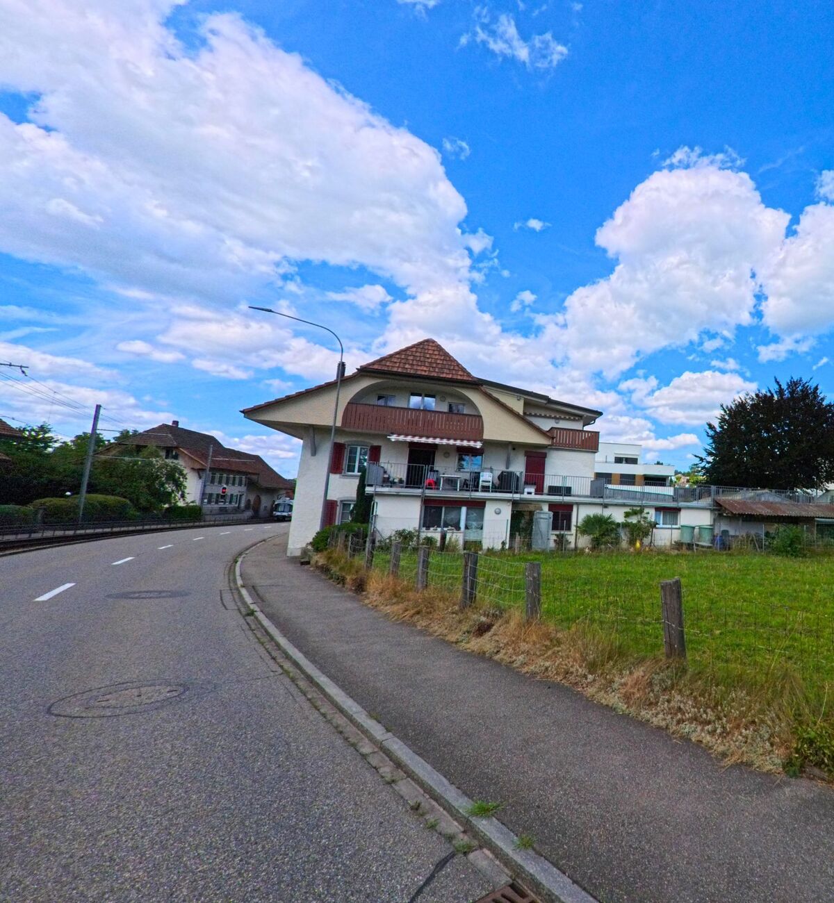 In Hirschthal (5042, Kanton Aargau) steht ein auffaelliges Wohnhaus mit geschwungener Fassade und rotem Ziegeldach direkt an einer leicht gekruemmten Hauptstrasse neben einer umzauenten Wiese.
