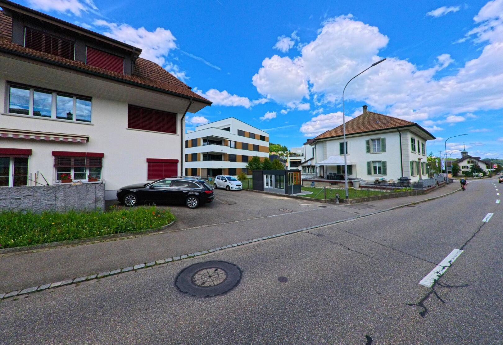 In Hirschthal (5042, Kanton Aargau) treffen traditionelle Wohnhaeuser mit Fensterlaeden auf moderne Neubauten entlang einer Hauptstrasse mit Bushaltestelle.