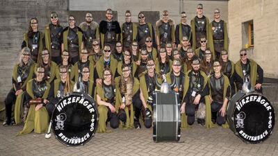 Eine grosse Fasnachtsgruppe in einheitlichen Kostuemen mit Trommeln steht zum Gruppenfoto in Breitenbach (4226, Kanton Solothurn) vor einer Betonwand.