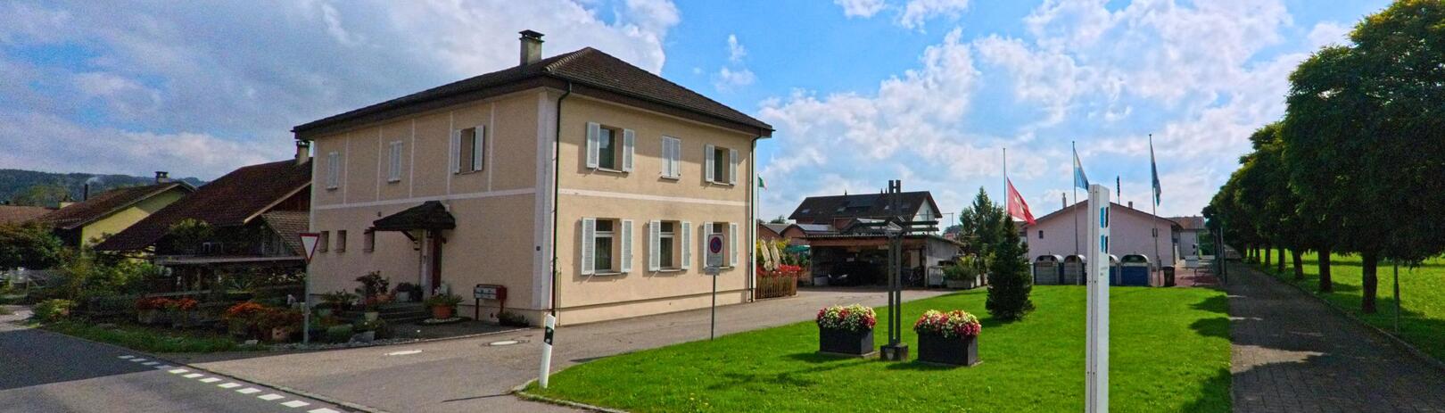 In Hitzkirch (6295, Kanton Luzern) steht ein helles Wohnhaus an einer kleinen Strasse, flankiert von gepflegten Gärten und einem Weg mit Bäumen.