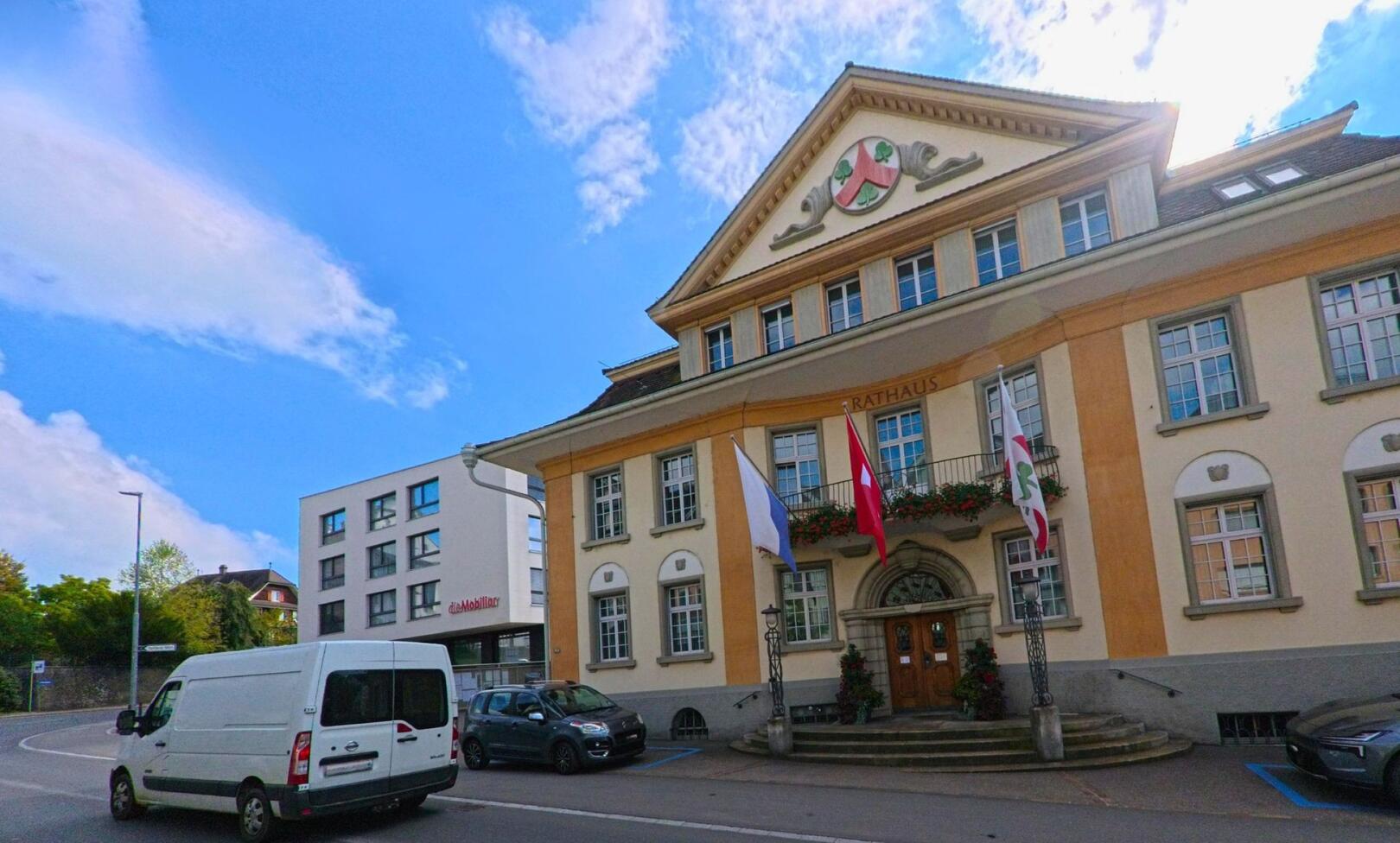 Das imposante Rathaus von Hochdorf (6280, Kanton Luzern) mit einer klassischen Architektur und farbenfrohen Blumenkästen an den Fenstern, vor einem modernen Gebäude.