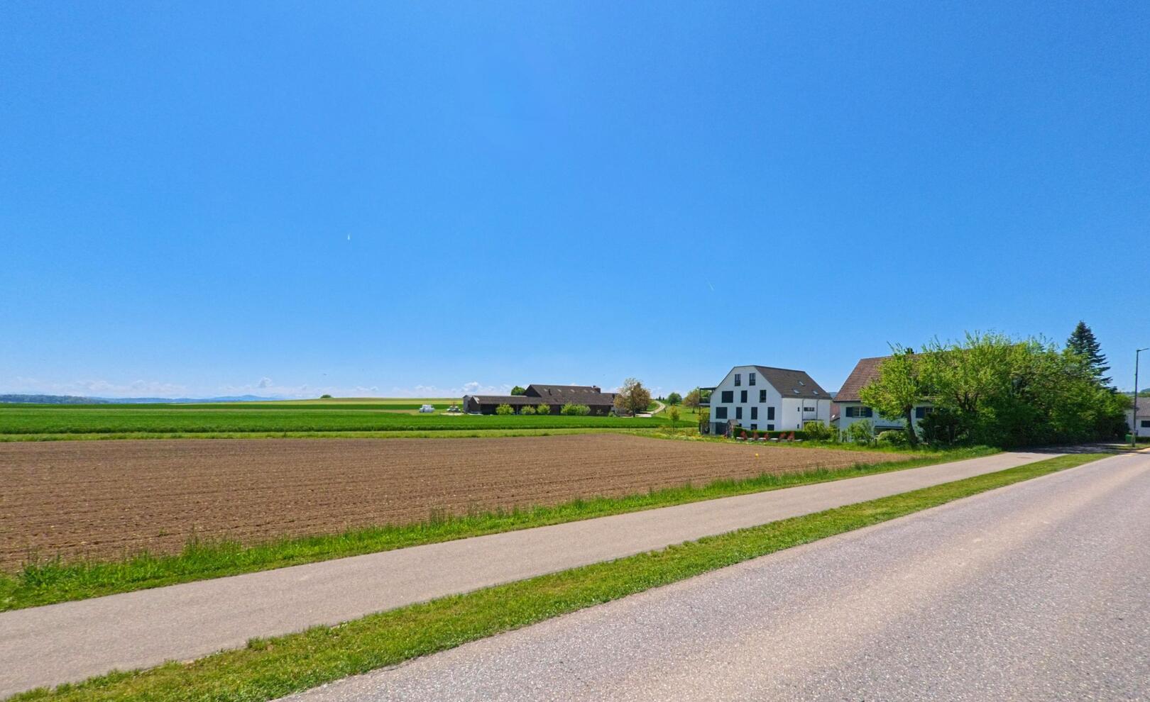 In Hochfelden (8182, Kanton Zürich) verläuft eine asphaltierte Strasse entlang von Feldern und einem Bauernhof, während sich der weite Himmel über die ländliche Landschaft erstreckt.