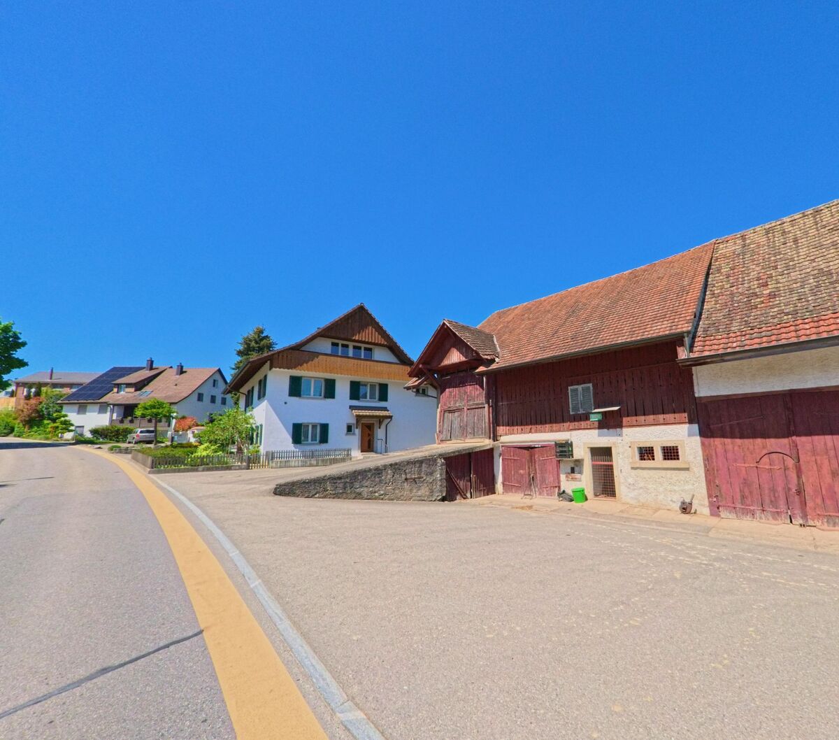 In Hochfelden im Kanton Zürich (PLZ 8182) verläuft eine kurvige Strasse an einem historischen Bauernhaus mit Scheune und mehreren Wohnhäusern entlang.