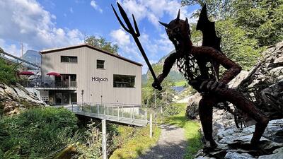 Vor dem Hölloch Center in Muotathal (6436, Kanton Schwyz) steht eine Teufelsstatue mit Dreizack, während das moderne Besuchergebäude im Hintergrund liegt.