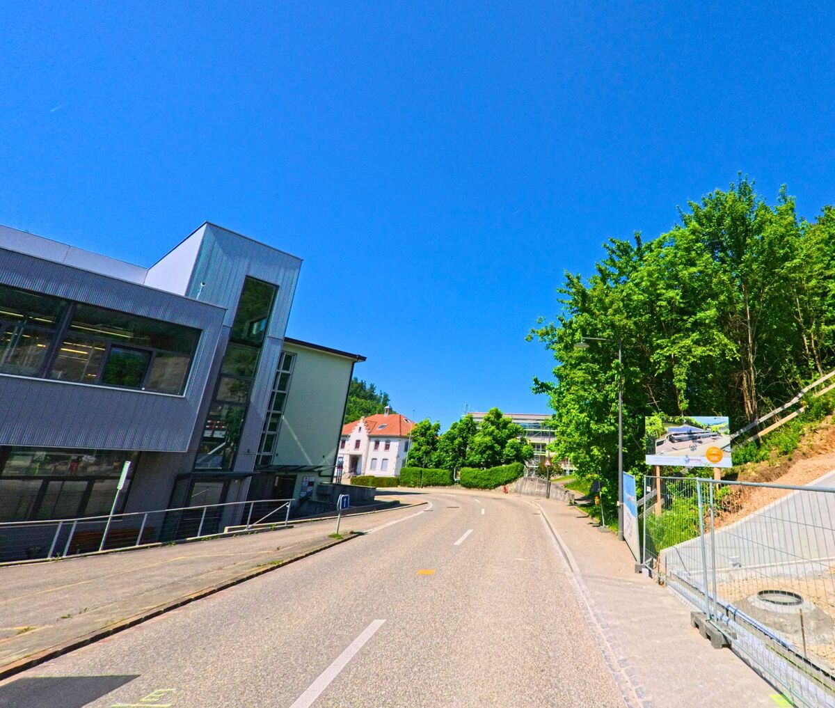 Eine Strasse in Hölstein (4434, Kanton Baselland) führt an einem modernen Schulgebäude vorbei, während auf der gegenüberliegenden Seite Bauarbeiten an einem Hang stattfinden.