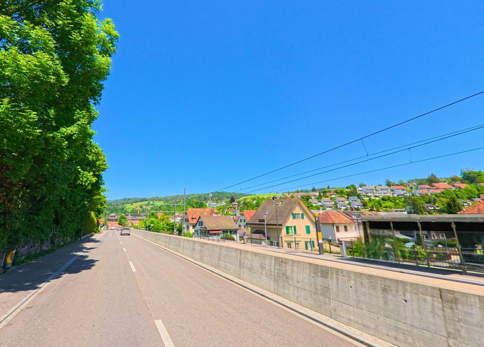 In Hölstein (4434, Kanton Baselland) verläuft eine breite Strasse parallel zu den Bahngleisen mit Blick auf Wohnhäuser und bewaldete Hügel im Hintergrund.
