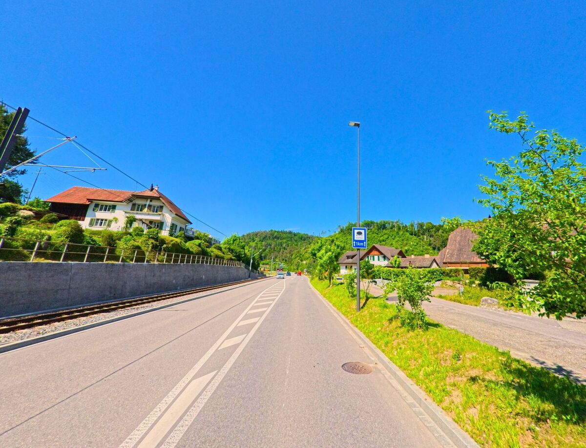 In Hölstein (4434, Kanton Baselland) verläuft eine Strasse mit Busstation parallel zu Bahngleisen, flankiert von Wohnhäusern und gepflegten Gärten.