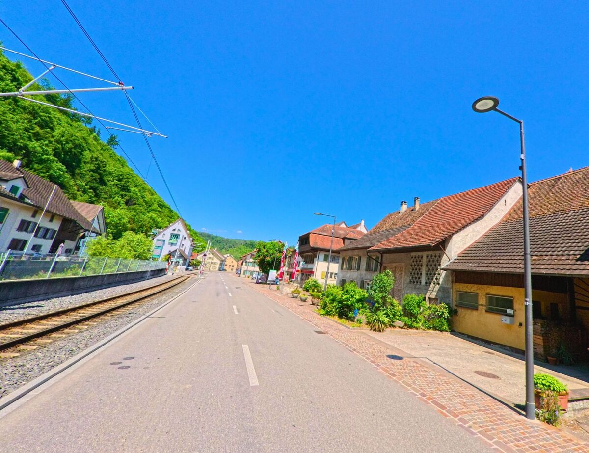 In Hölstein (4434, Kanton Baselland) führt eine Strasse mit Bahngleisen auf der linken Seite vorbei an traditionellen Häusern mit roten Ziegeldächern und Vorgärten.