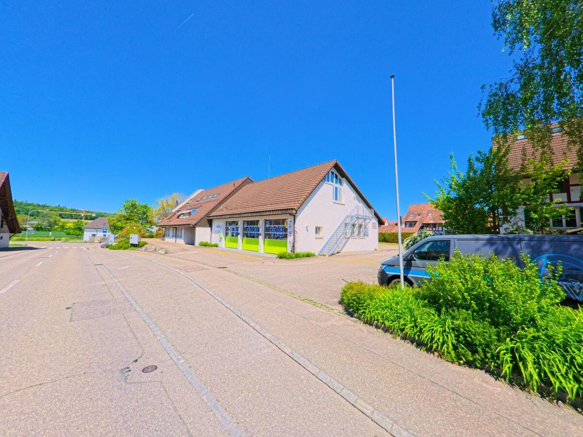 In Höri im Kanton Zürich mit der PLZ 8181 steht ein Gebäude mit markanten grünen Toren entlang einer breiten Strasse, umgeben von Wohnhäusern und leicht hügeliger Landschaft im Hintergrund.