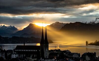 Die Kirche St. Leodegar im Hof in Luzern (6005, Kanton Luzern) steht im Vordergrund, während die Sonne hinter den Bergen golden über dem See aufgeht.